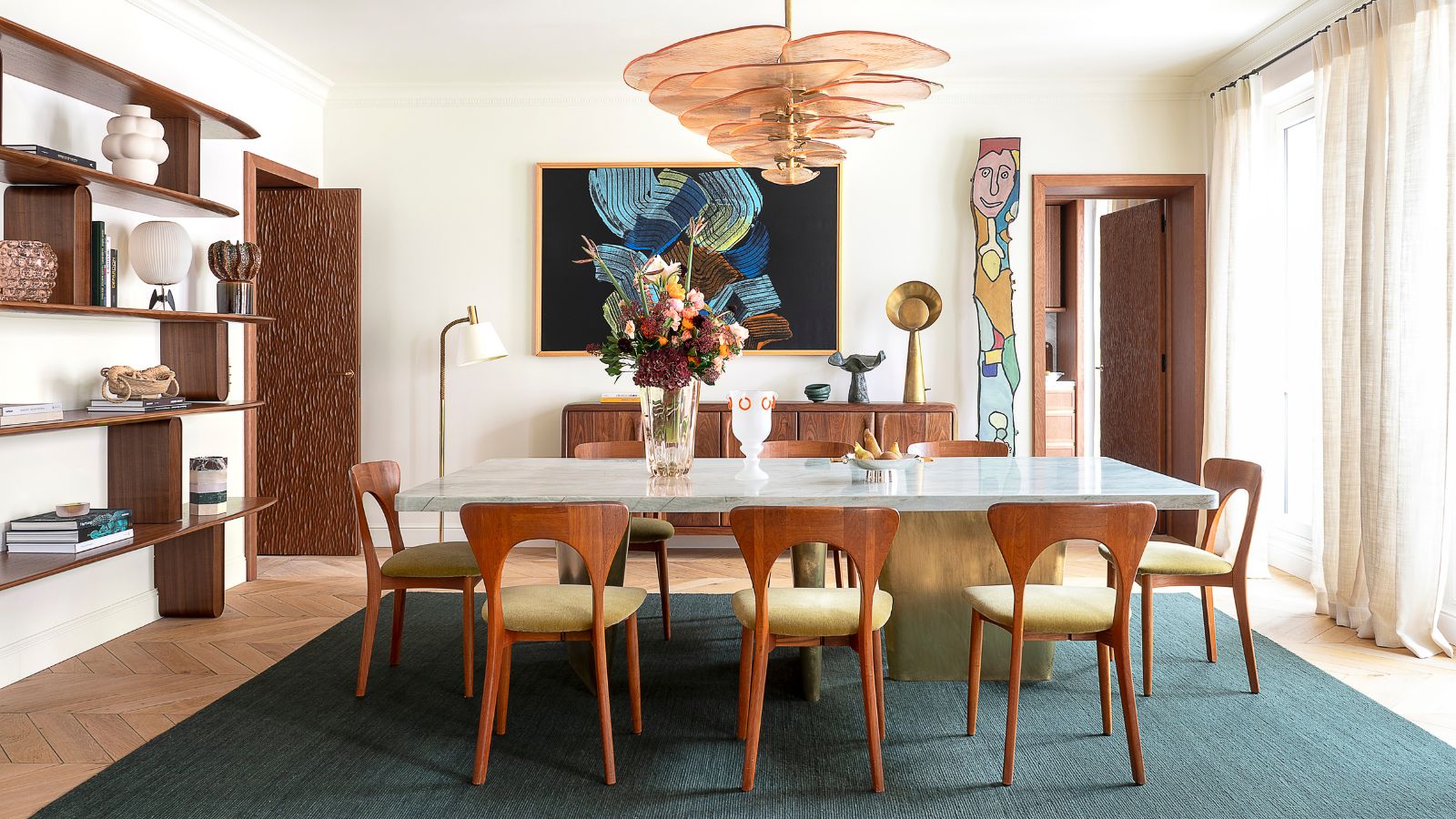 A spacious dining toom with a white rectangular table surrounded by modern wooden chairs. A large pink glass ceiling light hangs above. Mid-century modern shelving on the wall to the left. 