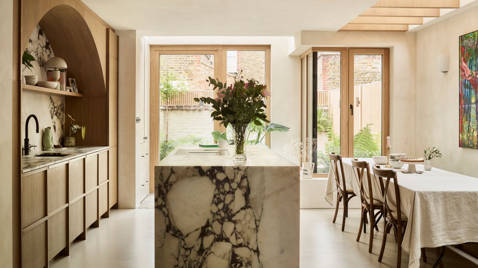 An airy kitchen diner with a large marble island separating worksurfaces and a rectangular dining table with three chairs pulled up to it. 