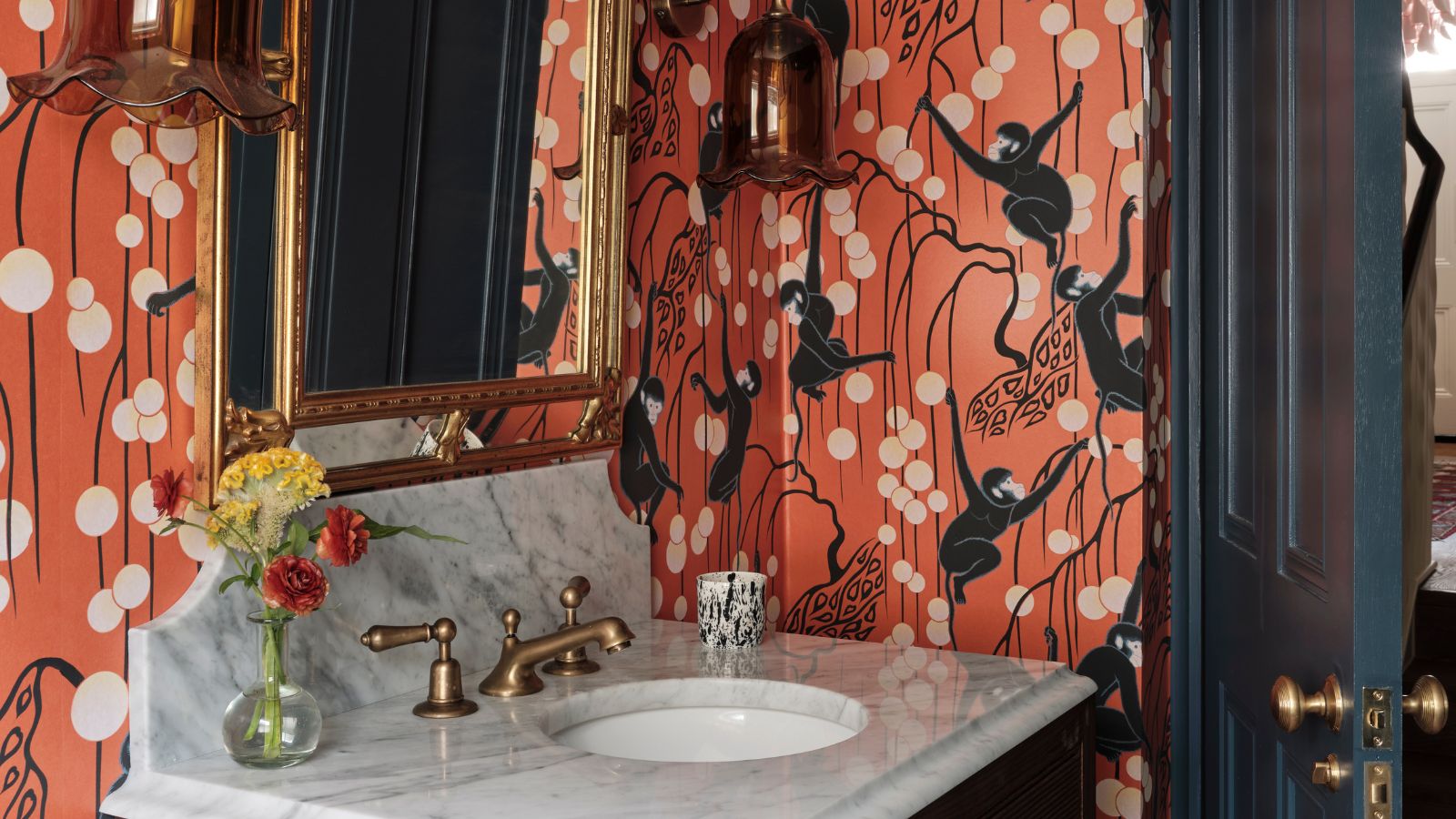 A small marble sink and backsplash in a small bathroom with terracotta wallpaper showing stripes, round white anterns and monkeys. The hardware and mirror is brass and the wooden door is painted dark blue