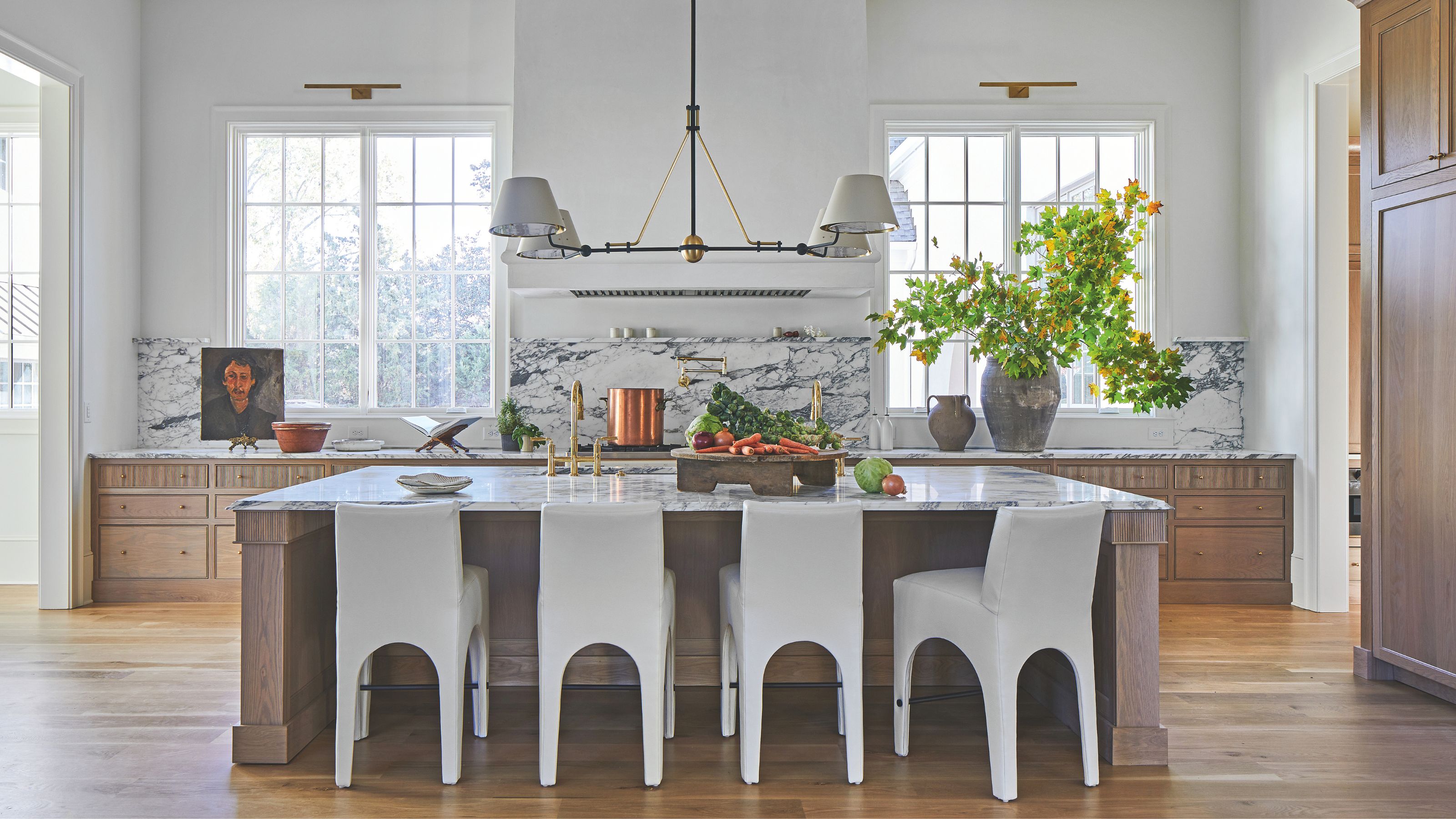 A large airy kitchen with white walls and wooden cabinets. The is a large kitchen island in the center with white chairs and a pendant light hanging above