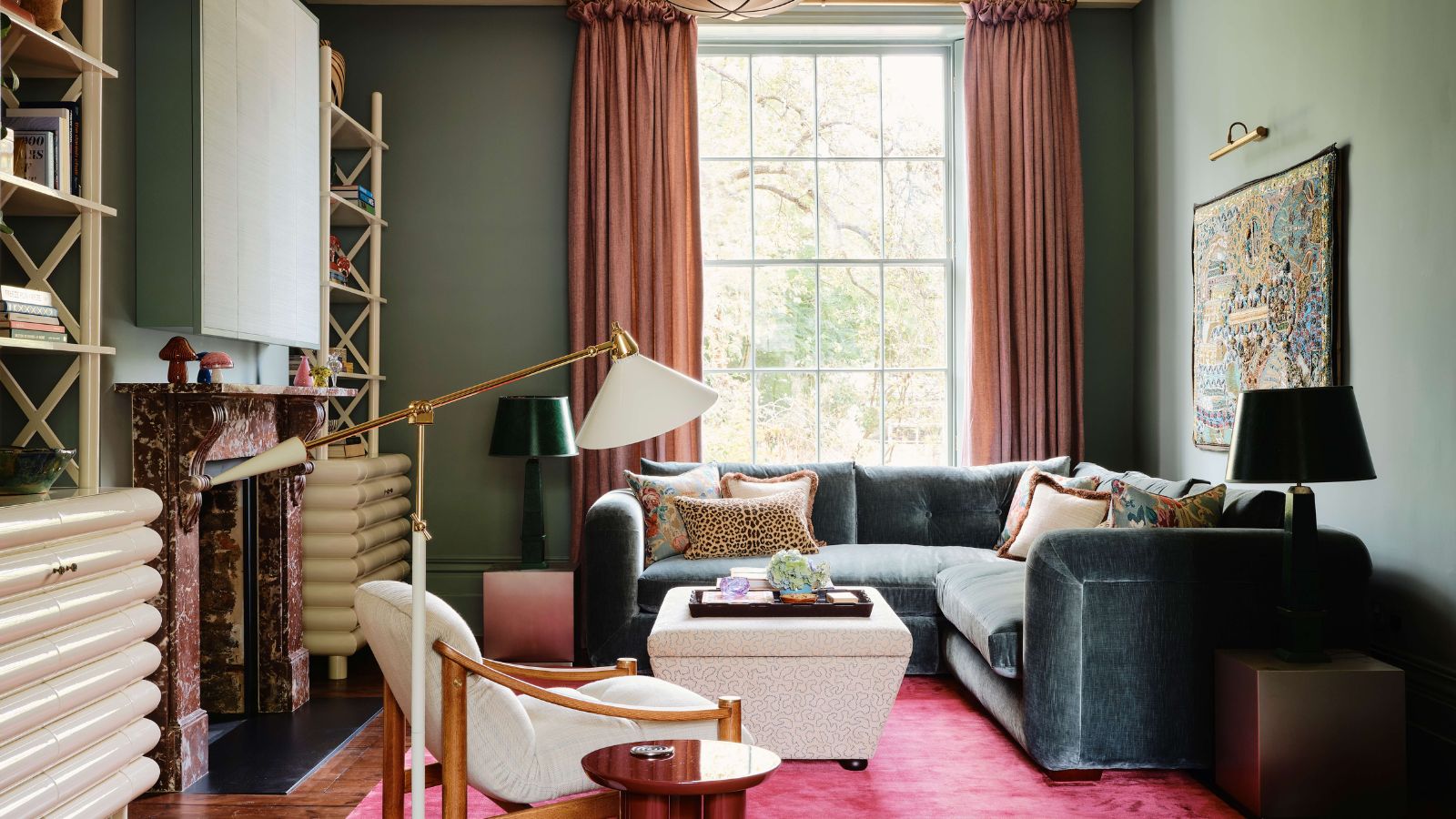A living room with teal walls, a bright pink rug, and a teal velvet corner sofa in front of a large window. 