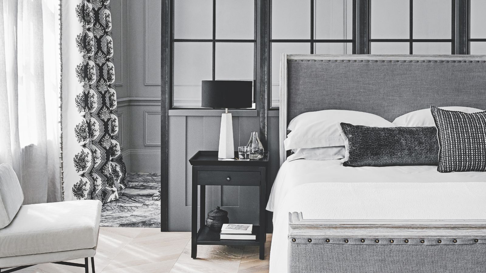 A gray bedroom with a large gray upholstered bed to the right, with white bedding and gray cushions. Beside the bedside table, to the left, is a black bedside table with books, vases, and a white lamp with a black lampshade. To the far right is a sheer white curtain over a window, and a white chair. The walls are gray paneled.