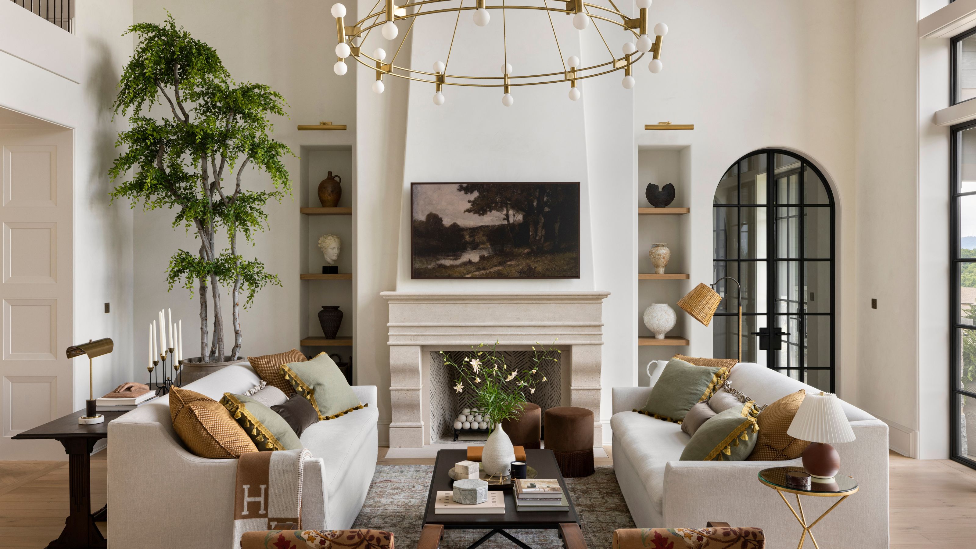 A bright living room with two white sofas facing a stone fireplace. A large tree sits in the corner, and shelves with pottery line the fireplace. A round gold chandelier hangs above, and tall black-framed windows let in natural light. A coffee table with books and flowers sits in the center.
