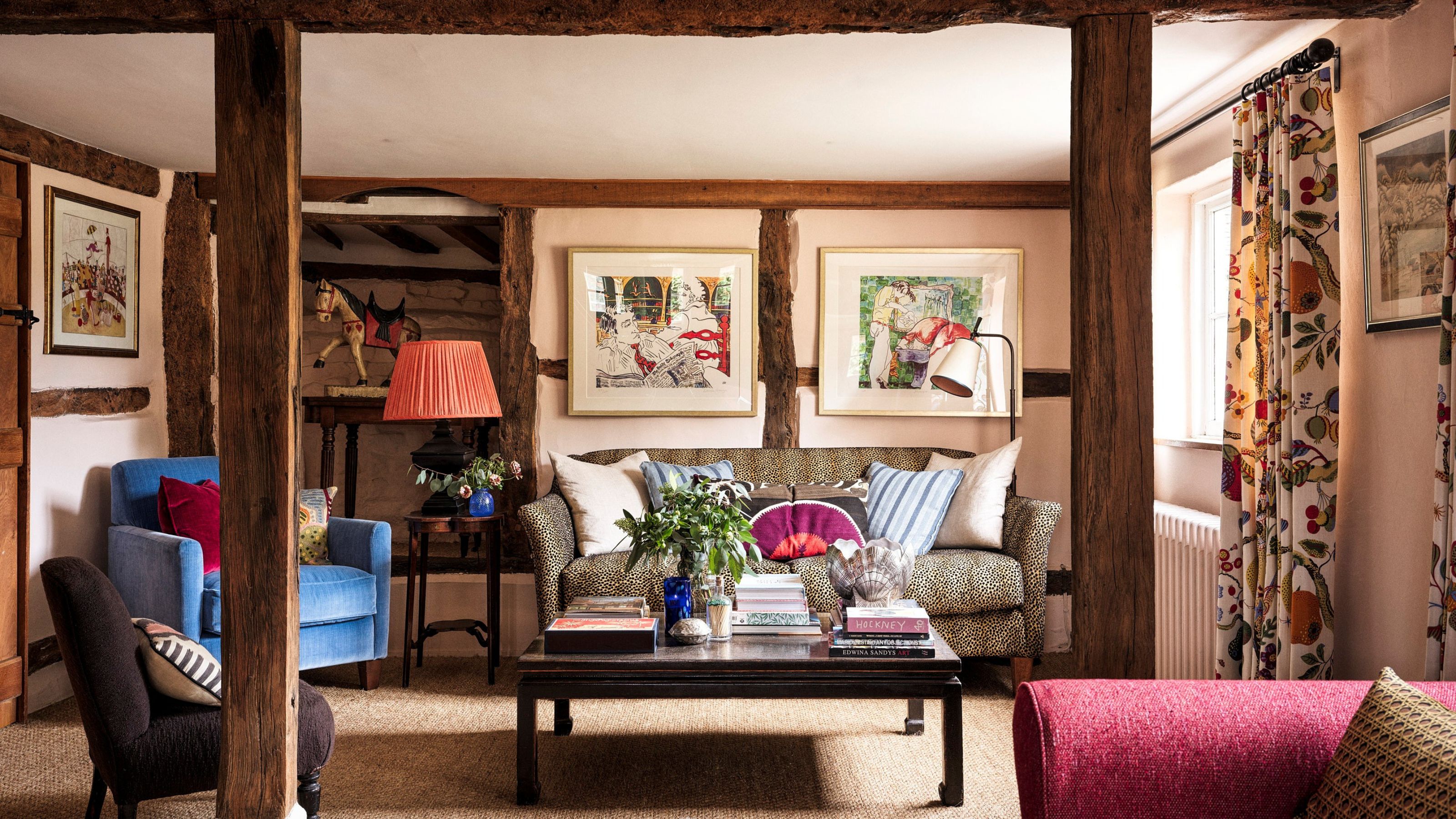 A cozy, rustic living room with exposed dark wood beams and pale pink walls. A leopard-print sofa with patterned cushions is centered around a dark wood coffee table stacked with books. Two framed artworks hang above the sofa. A bright red lamp and blue armchairs add pops of color