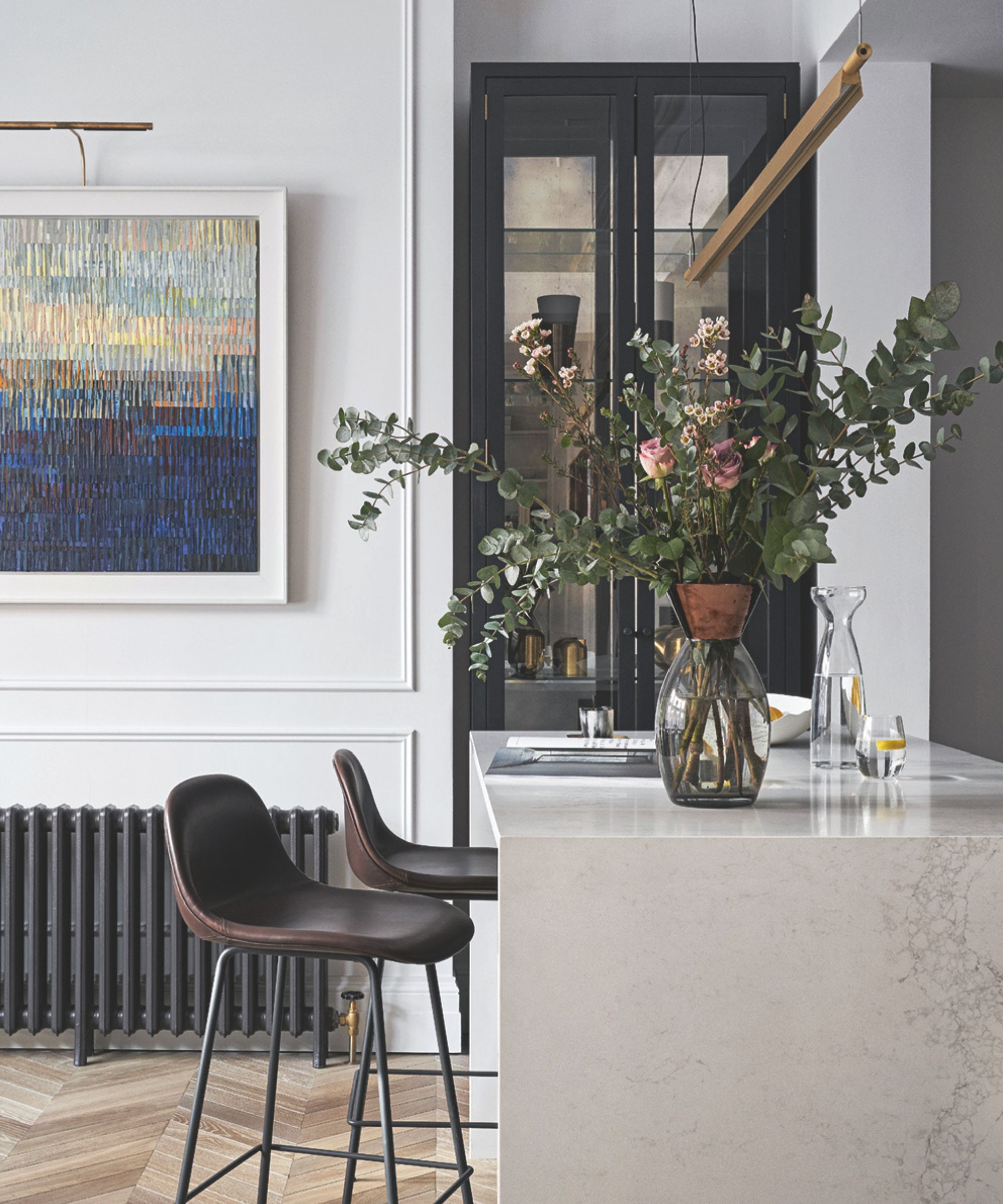 A white and modern kitchen space with a marble island, leather bar stools, wooden flooring, a glass vase filled with flowers, a black glass-fronted cabinet and a large abstract artwork on the wall.