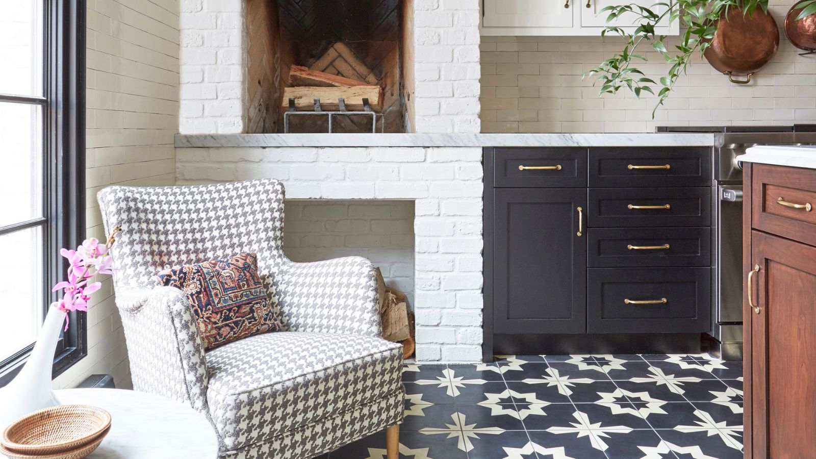 A kitchen corner with a white brick fireplace styled with an antique armchair reupholstered in a dogtooth fabric and a marble side table topped with a bud vase and pink flower