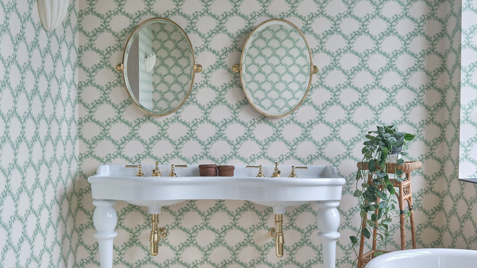 A his and her bathroom sink with two ovel mirrors above each sink. There is pretty green and white wallpaper on all of the walls and a rattan stand with a trailing plant sitting on top. 