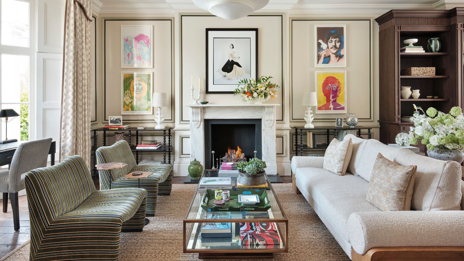 Luxe cream living room with hardwood floors and rug with paneled walls and bookshelves. There is a cream teddy four-seater sofa, and two single striped green chairs around a brass and glass coffee table. Floor-to-ceiling cabinets are built in, along with full height curtains, giving it a luxe look