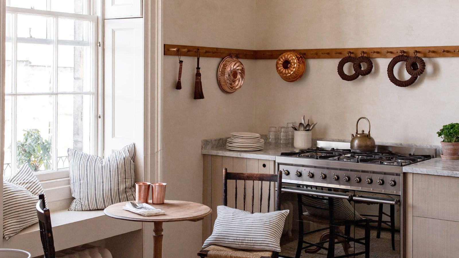 A warm neutral kitchen with wooden wall pegs displaying copper molds and a cozy eat-in area below the window with a small banquette, a round table, and two wooden chairs