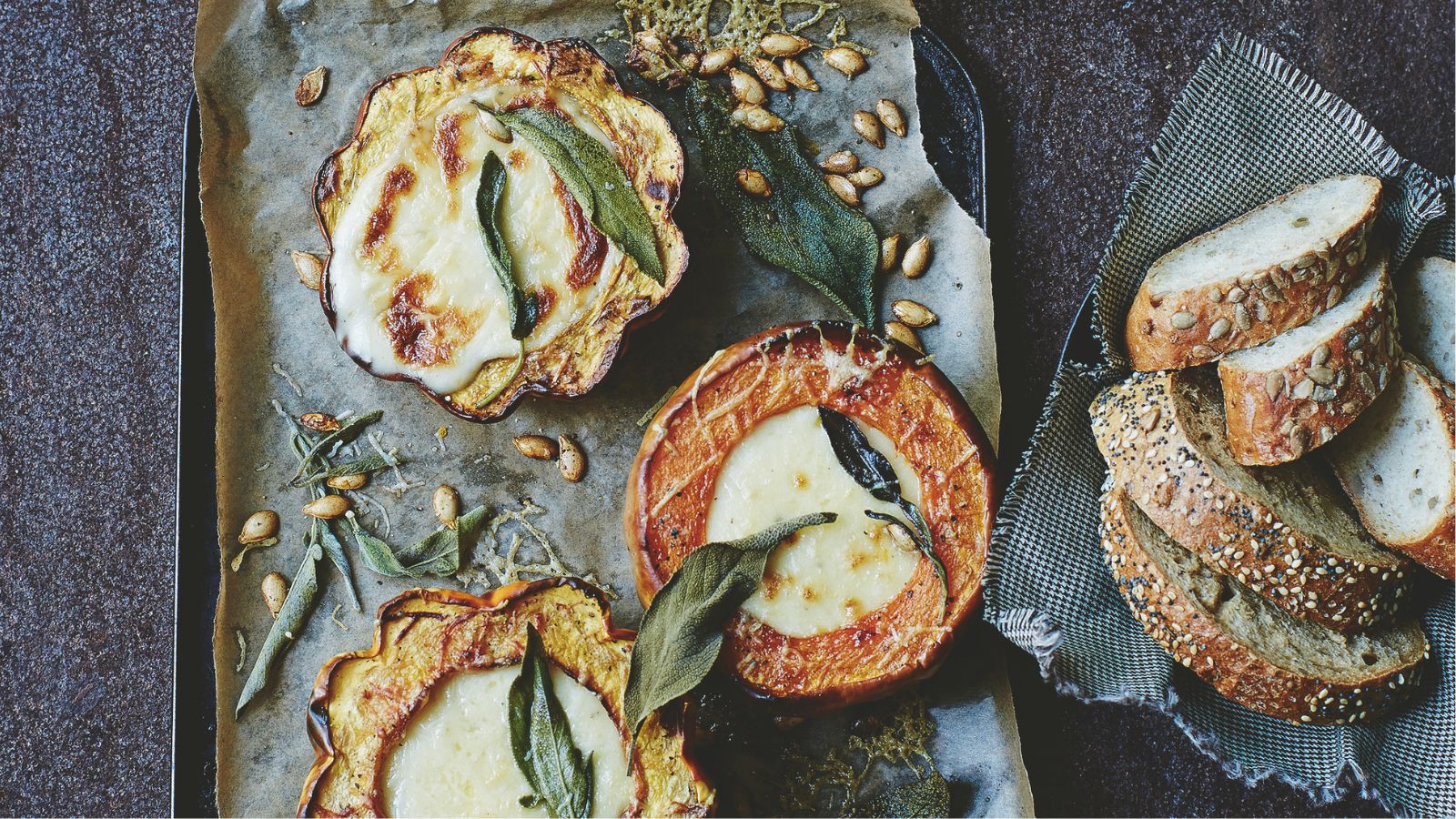 mini roast pumpkins filled with cheese fondue on a baking tray next to some seeded bread slices