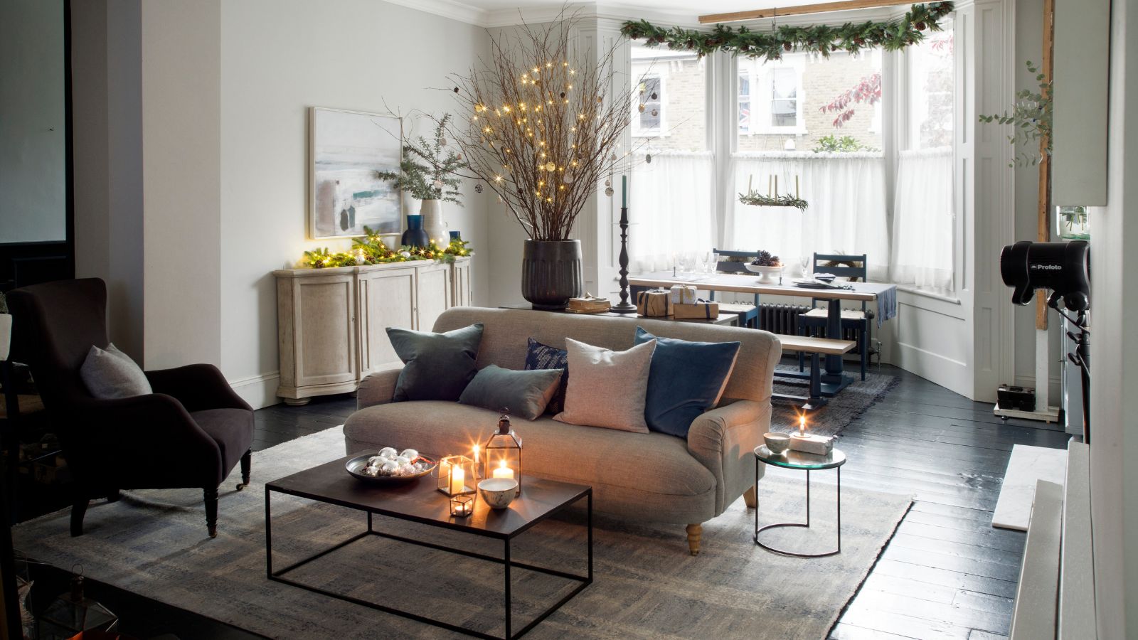 A cozy festive living room with a large cream sofa, coffee table with lit candles, large rug, brown upholstered chair, and a dining table under large bay windows with white sheer curtains. In the centre of the room is a large vase with lit dry twigs, and a tall candlestick. Above the window, festive greenery is hanging, and to the right is a white side-table with fairy lights, a vase of festive sprigs, and an artwork above.