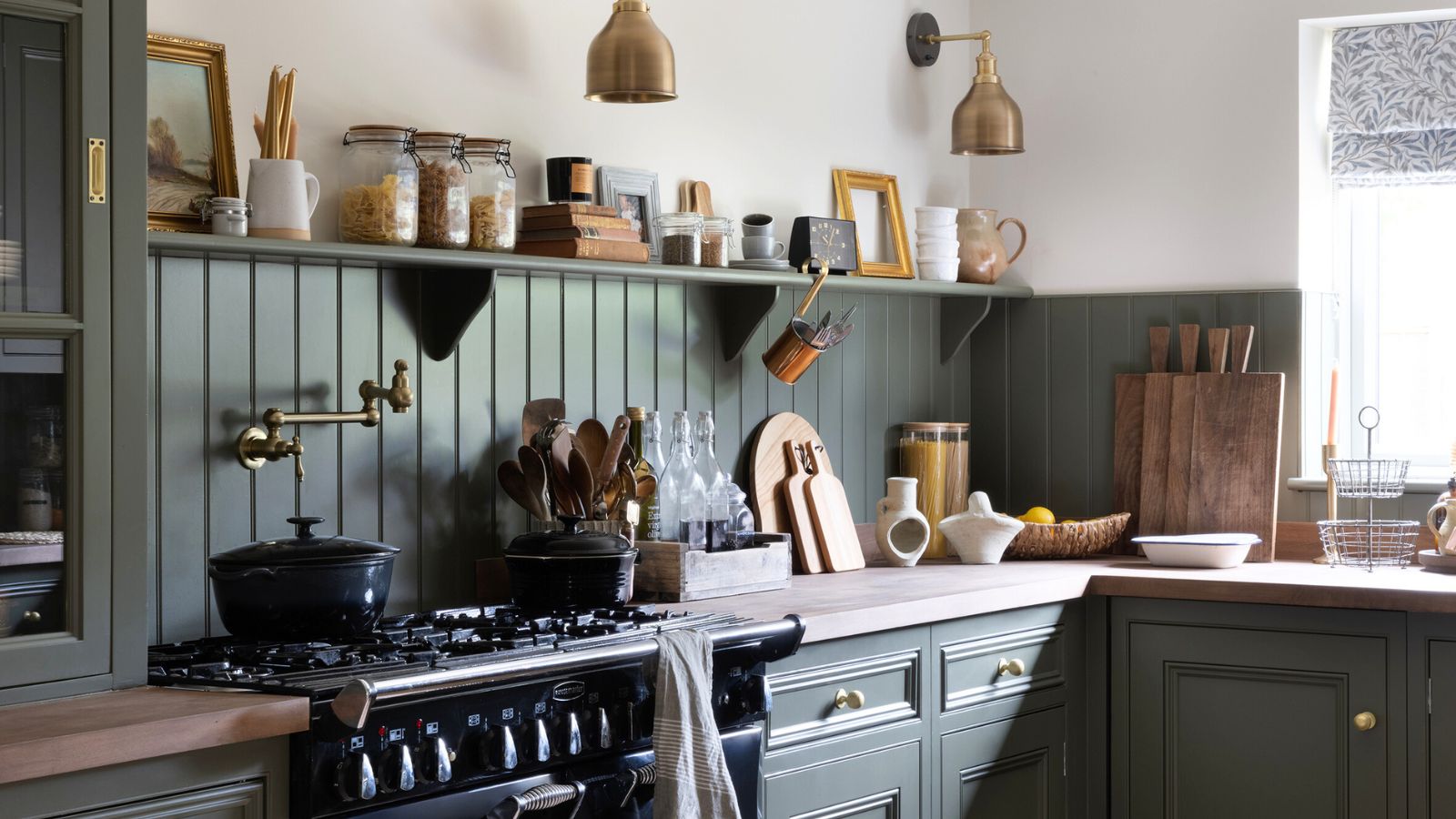 The best cookware in a rustic kitchen with green panelled backsplashes and matching cabinets, an old gas stove, and kitchen decor on shelving. 