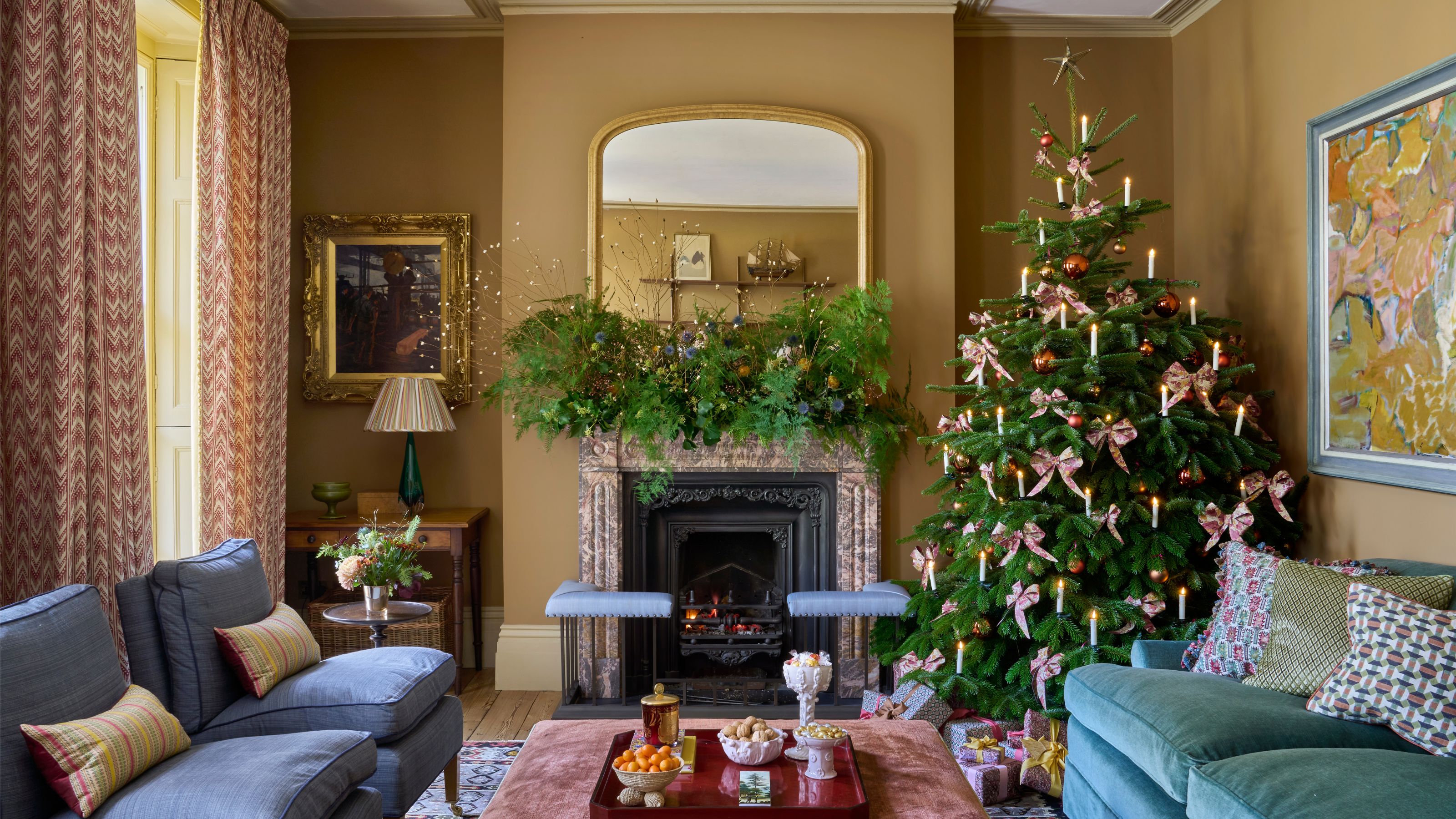 Cozy, traditional living room with mustard-yellow/brown walls. A large Christmas tree decorated with pink bows stands next to a fireplace mantle adorned with greenery. The seating includes a navy-blue sofa and a teal armchair.