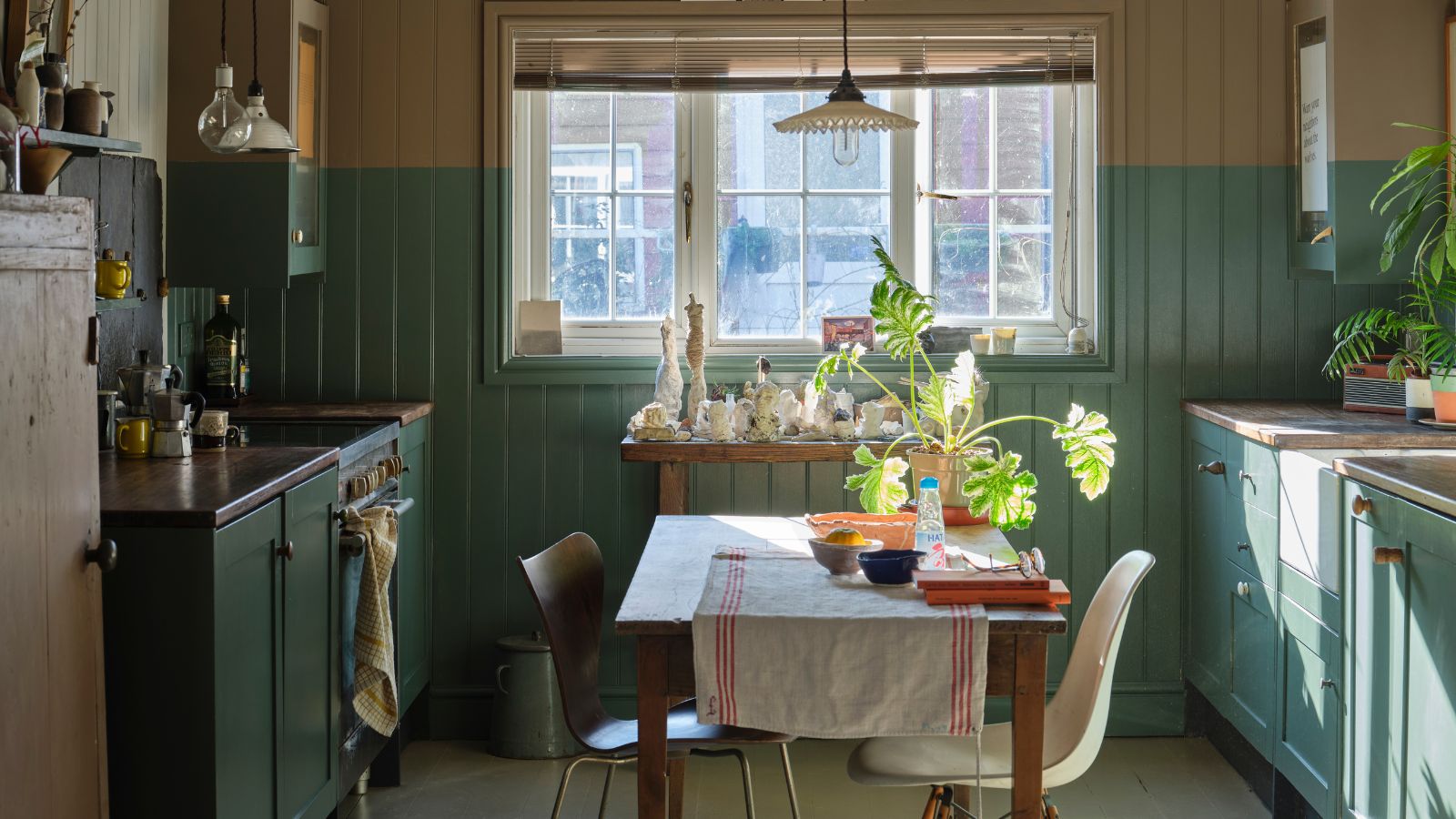 Small kitchen with sage green cabinets on left and right, with window at middle back, and table in middle fo space with two mismatched chairs.